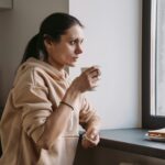 Mujer mirando por la ventana con taza de café y expresión cansada