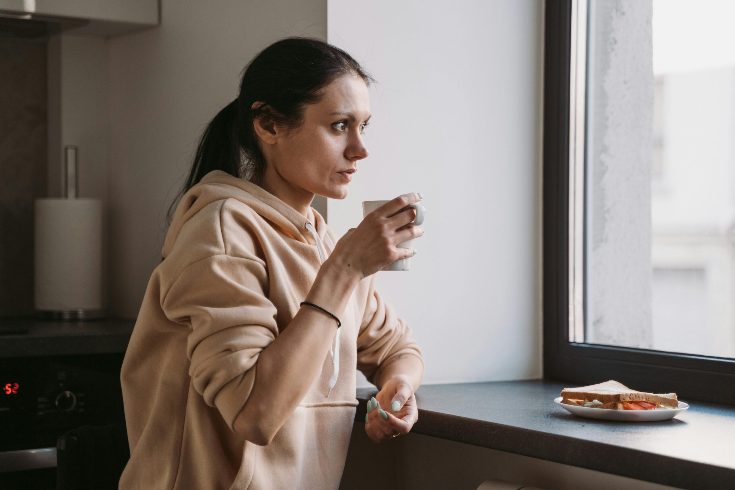 Mujer mirando por la ventana con taza de café y expresión cansada