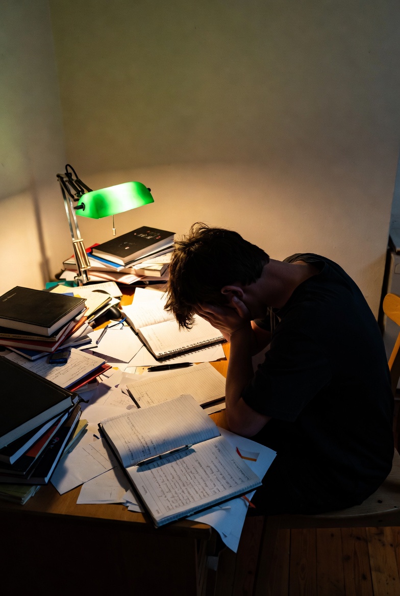 Estudiante adolescente frente a escritorio con apuntes, cabeza entre manos, expresión de agobio.